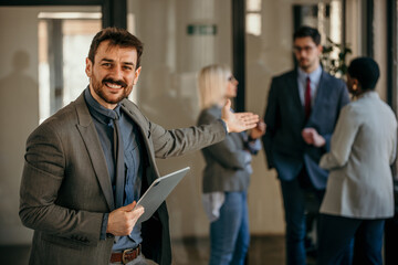 Portrait of smiling confident businessman in boardroom holding digital tablet and pointing to his team. A diverse group of businesspeople in meetings and talk behind the manager.