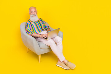 Full body photo of cheerful nice aged man sit armchair hold netbook empty space isolated on yellow color background