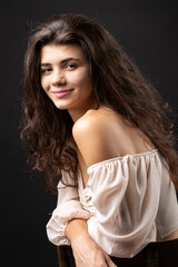Obraz premium Classic studio portrait of a young brunette in a white loose top, who is sitting on a chair against a black background.