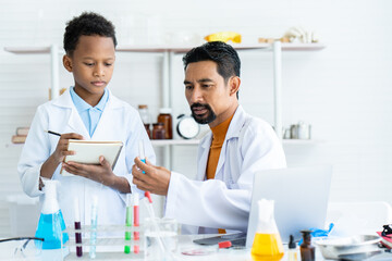 In chemistry classroom with many laboratory tools on table. A young African boy and male teacher in white lab coat doing experiment together. A boy writing data and teacher checking at blue test tube.