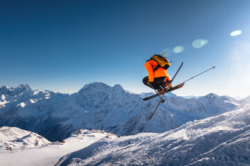 Naklejka na ściany i meble The skier jumps on the background of the blue sky and snow-capped mountains. freestyle skier performs helicopter with crossed skis simultaneously with full rotation