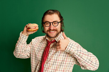 Emotive retro mature man with moustache and beard in eyeglasses in vintage fashion clothes tasting hamburger over dark green background.