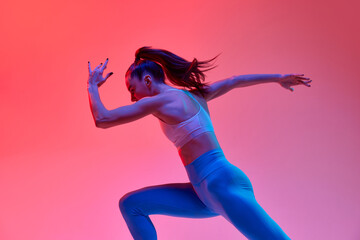 Young girl, professional runner athlete in uniform training, running over pink studio background in...
