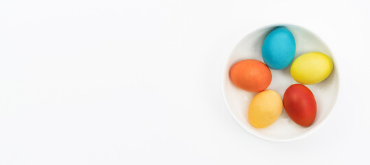 Easter eggs lie in a bowl on a white background with copy space. Easter banner in a minimalistic style. Traditional Easter eggs painted by kids in a white plate top view.