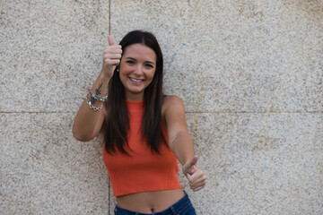 Beautiful brunette girl with long hair is happy and shows both hands with thumbs up because everything is correct and ok, isolated on beige background and orange top.