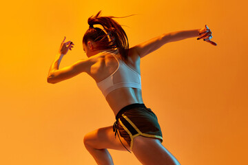 Young girl, professional runner athlete in uniform training, running over orange studio background in neon light. Concept of sportive lifestyle, health, endurance, action and motion. Ad