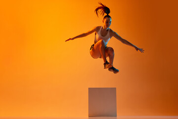 Workout. Young sportive girl, athlete training, jumping over block against orange studio background...