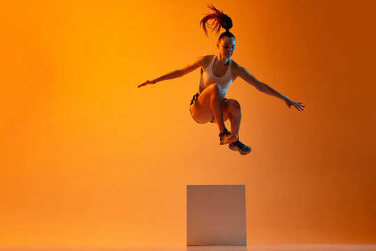 Workout. Young sportive girl, athlete training, jumping over block against orange studio background in neon light. Concept of sportive lifestyle, health, endurance, action and motion. Ad