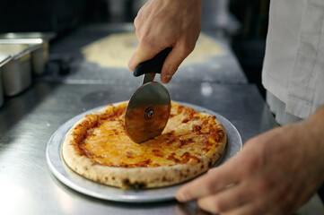 Chef cutting freshly baked pizza with round knife on wooden board
