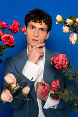 Curly man in stylish outfit touching chin near blooming flowers on blue background.