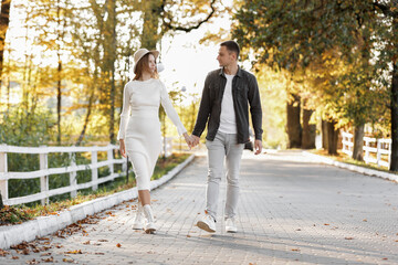 Fototapeta premium Pregnancy and Parenthoods concept - smiling man is walking with pregnant wife and looking to each other in autumn park. Happy styling young family is relaxing outdoors on weekend on sunny day.