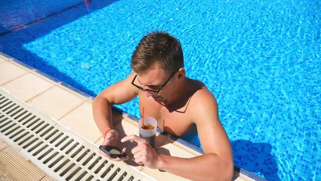 Handsome man using phone at pool of hotel and refreshing cold cocktail. Young guy in sunglasses drink beverage enjoying warm sunny day. Man relaxing on resort during summer vacation. Slow motion