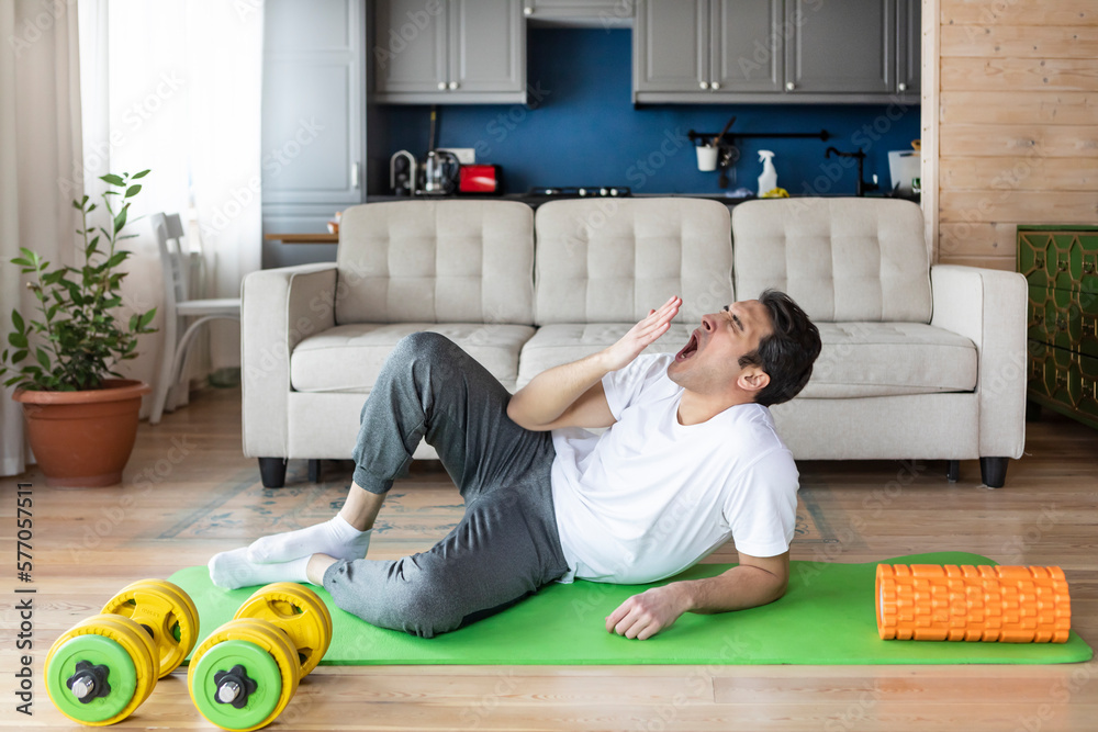 Wall mural tired young man yawning while training on the floor at home. do sport and exercise at the morning ti