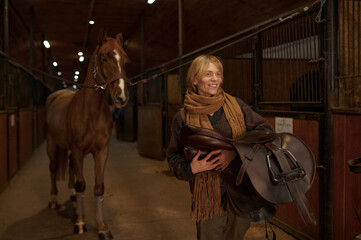 Female horse rider walking with harness in stable