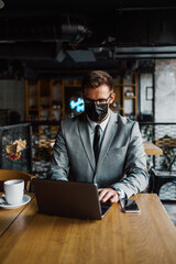 A businessman with a protective mask on his face is sitting in a cafe and talking on the phone while using laptop. Coronavirus, Covid-19 concept.