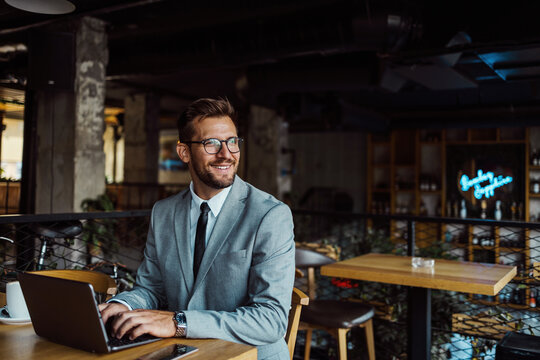 Middle age businessman sitting in cafe bar and enjoying at coffee break between two meetings. He uses laptop computer and he is positive and confident.