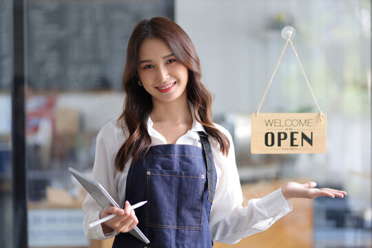 Portrait of a young female entrepreneur hanging a welcome sign in front of a coffee shop. Beautiful waitress or hostess holding a tablet preparing to advise food orders in a restaurant.