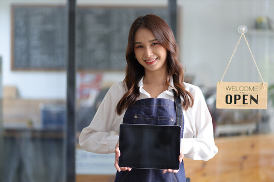 Portrait of a young female entrepreneur hanging a welcome sign in front of a coffee shop. Beautiful waitress or hostess holding a tablet preparing to advise food orders in a restaurant.