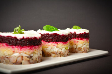 Side view of traditional layered fish salad with beetroot, potato, onion, herring and mayonnaise. Salad herring under fur coat served in culinary rings in focus and out of focus on dark background