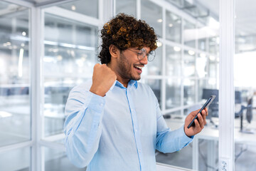 Successful hispanic programmer inside office and celebrating victory and successful achievement, man reading received news online phone, businessman in shirt holding hand up triumph gesture