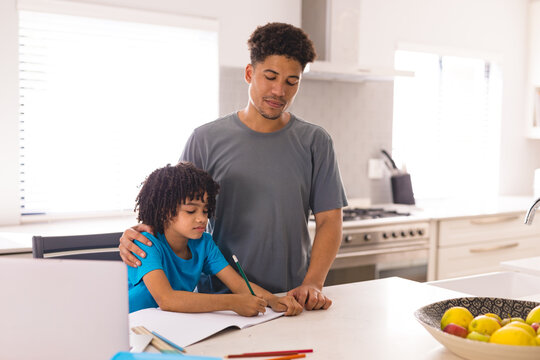 Hispanic man standing by son doing homework at dining table in kitchen