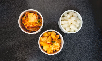 Obraz na płótnie Canvas Three traditional kimchi types in white bowls (turnip, spicy turnip and lettuce) in table in kitchen with copy space (kimchi: pickled and fermented vegetables, garlic, ginger, chili peppers, etc)