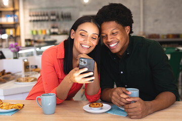 Happy young multiracial couple sharing smartphone while sitting with coffee together in cafe