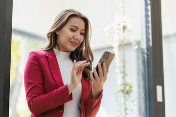 Asian female worker sitting in remote office holding smartphone using business applications or...