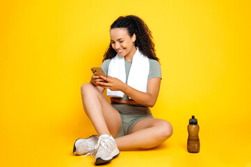 Full length photo of joyful lovely mixed race woman, in sportswear, with a towel on her shoulders, sits on isolated yellow background, using cellphone, messaging, watching online workout videos, smile