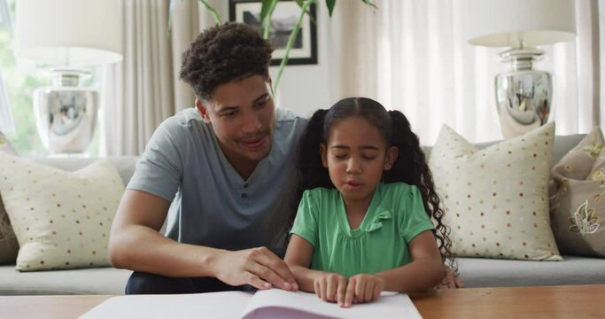Happy biracial father and daughter sitting on sofa reading braille