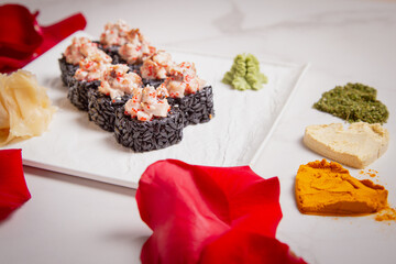 Side view of black rice sushi roll with Unagi eel. Red rose petals and heart shape spices near served dish on marble background. Sushi roll portion on white background