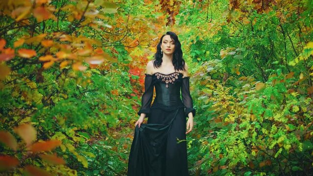 Queen fantasy woman in black gothic old style dress walks, autumn nature forest trees. Green yellow orange foliage park. princess girl sexy fashion model. royal vintage long dark ball gown. video 4k