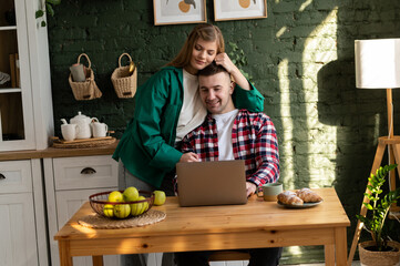 Young couple in the kitchen. A young woman hugs the head of her husband, who is sitting at a wooden table in front of a laptop and working. Horizontal.