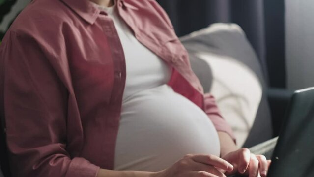Pregnant woman using laptop at home. Close-up of cute smiling young future mother with computer. Searching, online communication, shopping via Internet or working during pregnancy and maternity leave
