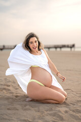 Vertical photo of exited pregnant woman in white dress sitting on the beach.