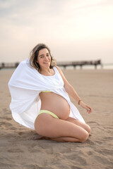 Full length portrait of pregnant young woman sitting on the beach and showing her tummy. Pregnancy, motherhood and baby expectation concept.