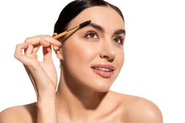 brunette woman styling eyebrows with brush while looking away isolated on white.