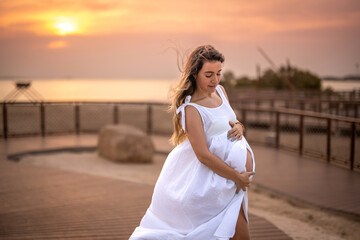 Fototapeta na wymiar 9 months pregnant woman wearing white dress and touching her belly on the beach walkway at sunset.