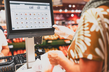 Senior woman wearing protective gloves weighs her purchases of vegetables on the electronic scale of the supermarket