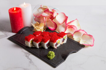 Obraz na płótnie Canvas Side view of Japanese sushi roll with tuna, avocado and flying fish roe on top. rose petals and candles near served dish. Sushi roll portion with ginger, wasabi on white marble background