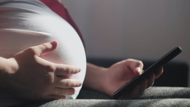 Close up of young pregnant woman in casual clothes sitting on cozy couch in light living room and using smartphone. Finger touching screen. Baby expectation time. Home relax and pregnancy concept