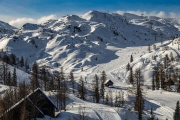 Bohinj, Slovenia - Winter view of the snowy mountain Vogel with skiers on ski slopes and wooden huts in the Alps at Triglav National Park on a sunny winter day with blue sky