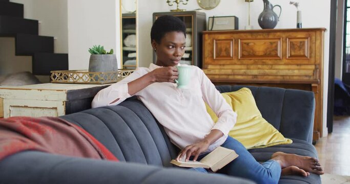 Happy african american woman sitting on sofa in living room, reading book