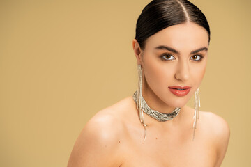 brunette young woman in necklace and earrings looking at camera isolated on beige.