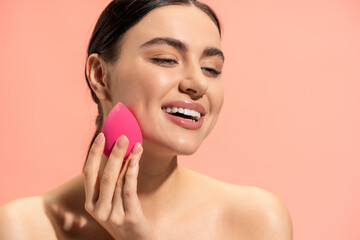 happy woman applying face foundation with beauty sponge isolated on pink.