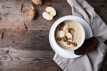 Bowl of homemade Jerusalem artichoke and apple cream soup with slice of bread and fresh sun choke tubers and apple on wooden table.