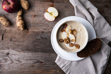 Bowl of homemade Jerusalem artichoke and apple cream soup with slice of bread and fresh sun choke tubers and apple on wooden table.