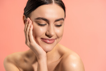 pleased woman with naked shoulders touching soft cheek while looking away isolated on pink.