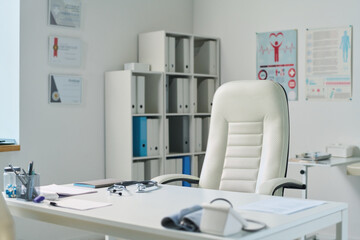 Empty medical office with workplace of clinician consisting of desk and white leather armchair standing against shelves with folders