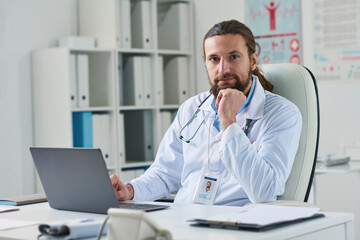 Young successful male clinician in lab coat sitting by workplace in front of laptop computer in medical office and looking at camera